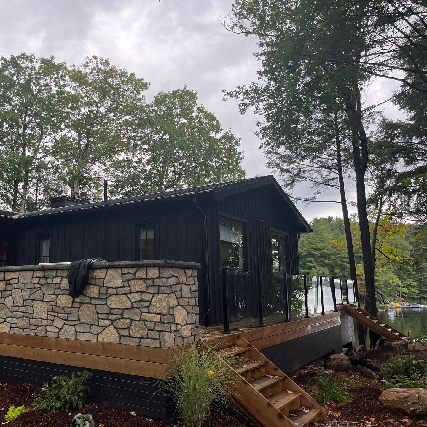Black cabin with stone base on a wooded lakeside, with wooden steps and deck under cloudy skies
