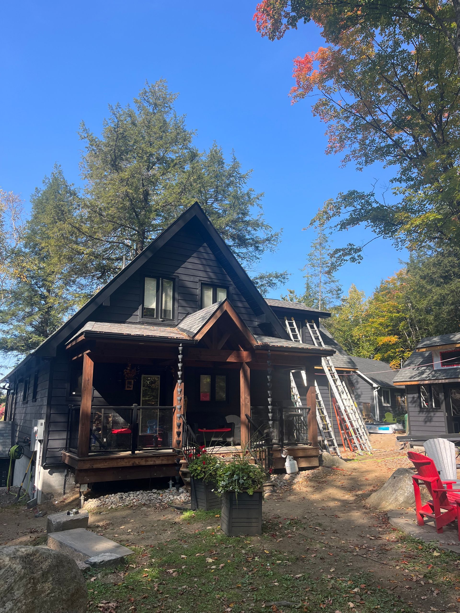Rustic cabin with a porch in a wooded yard, autumn trees and red chairs under a bright blue sky.