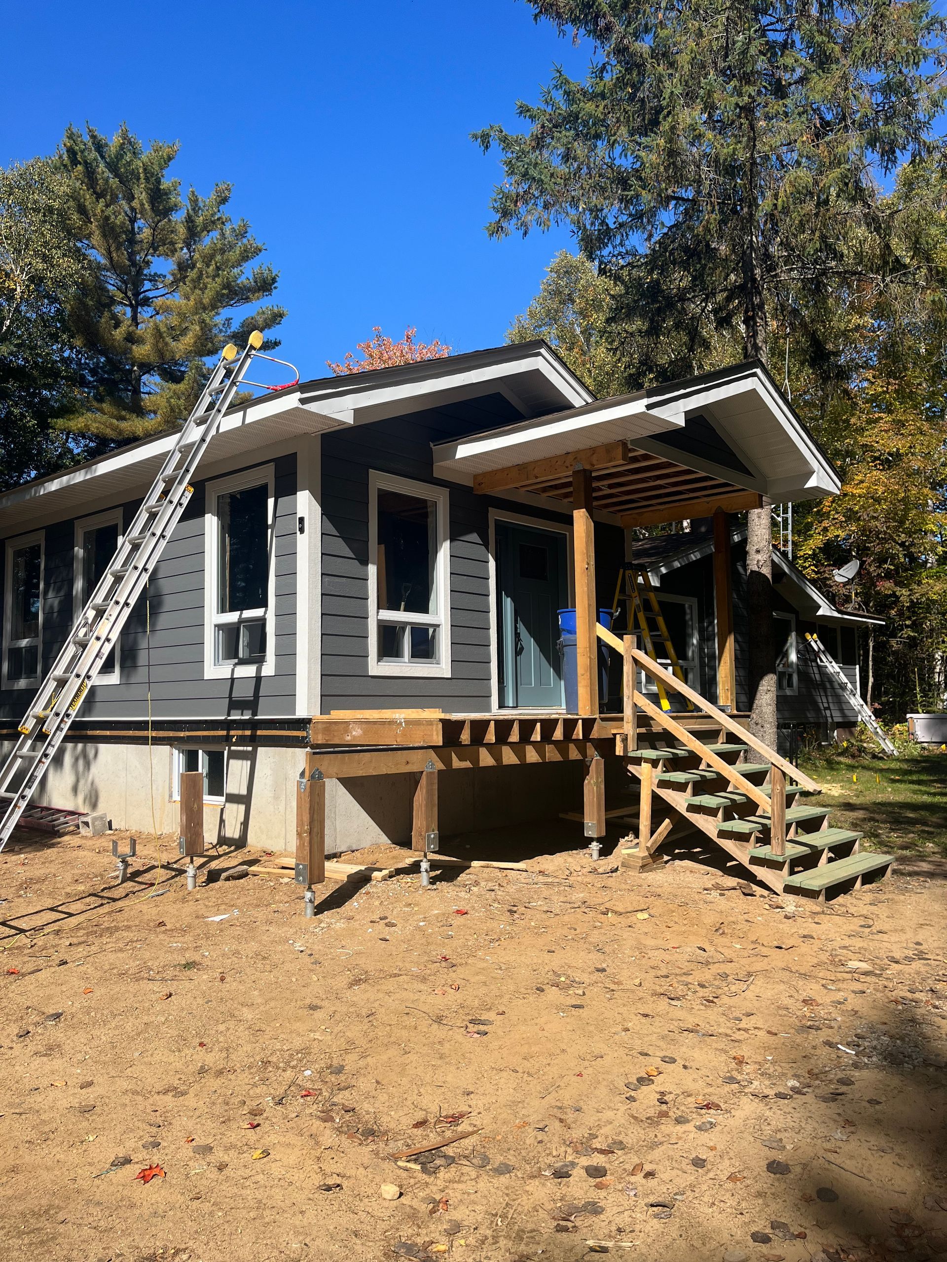 Gray house under renovation with front porch, stairs, ladder, and bare dirt yard under a blue sky