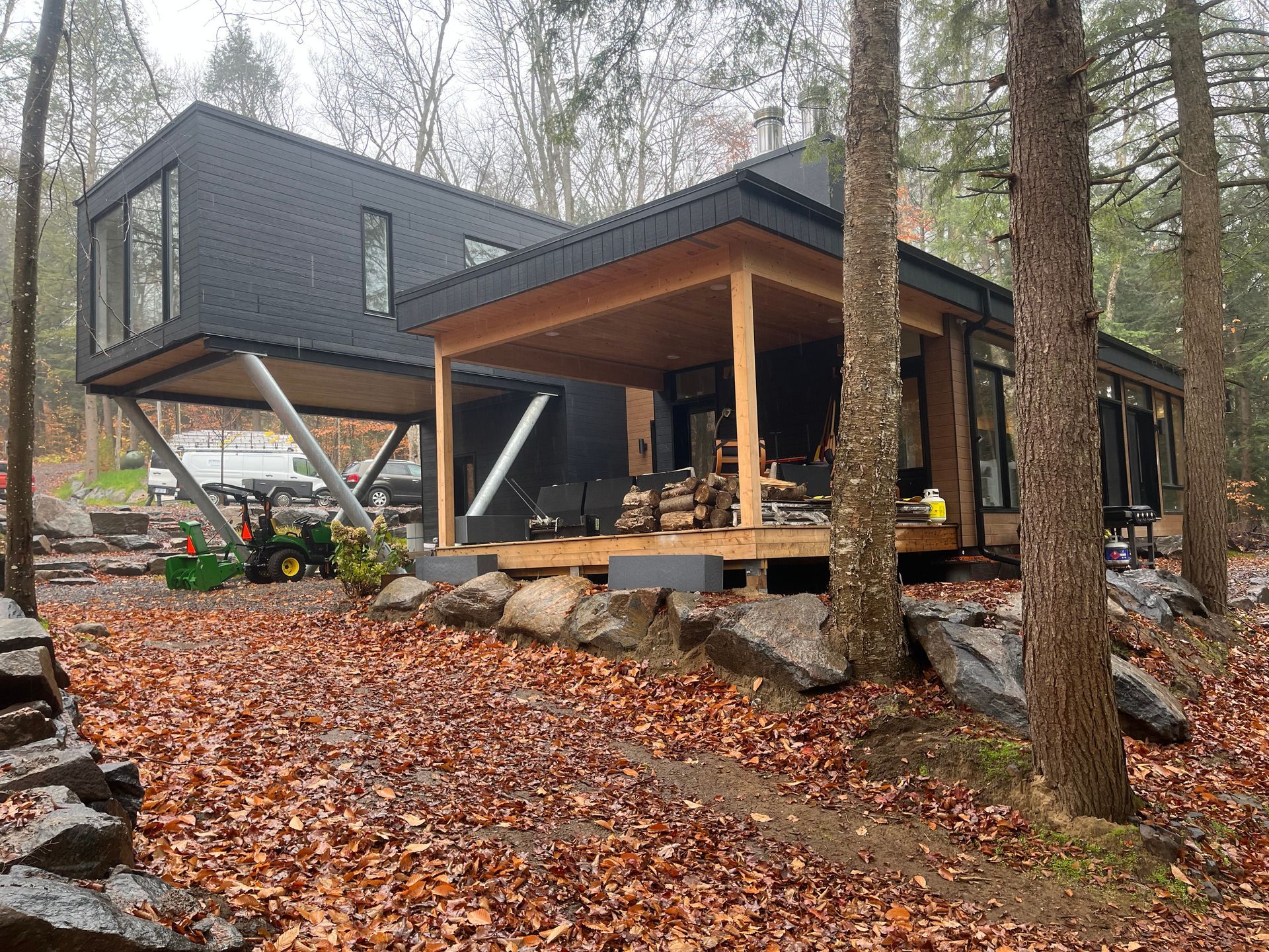 Modern cabin with elevated dark gray section and wood porch in a wooded, leaf-covered setting