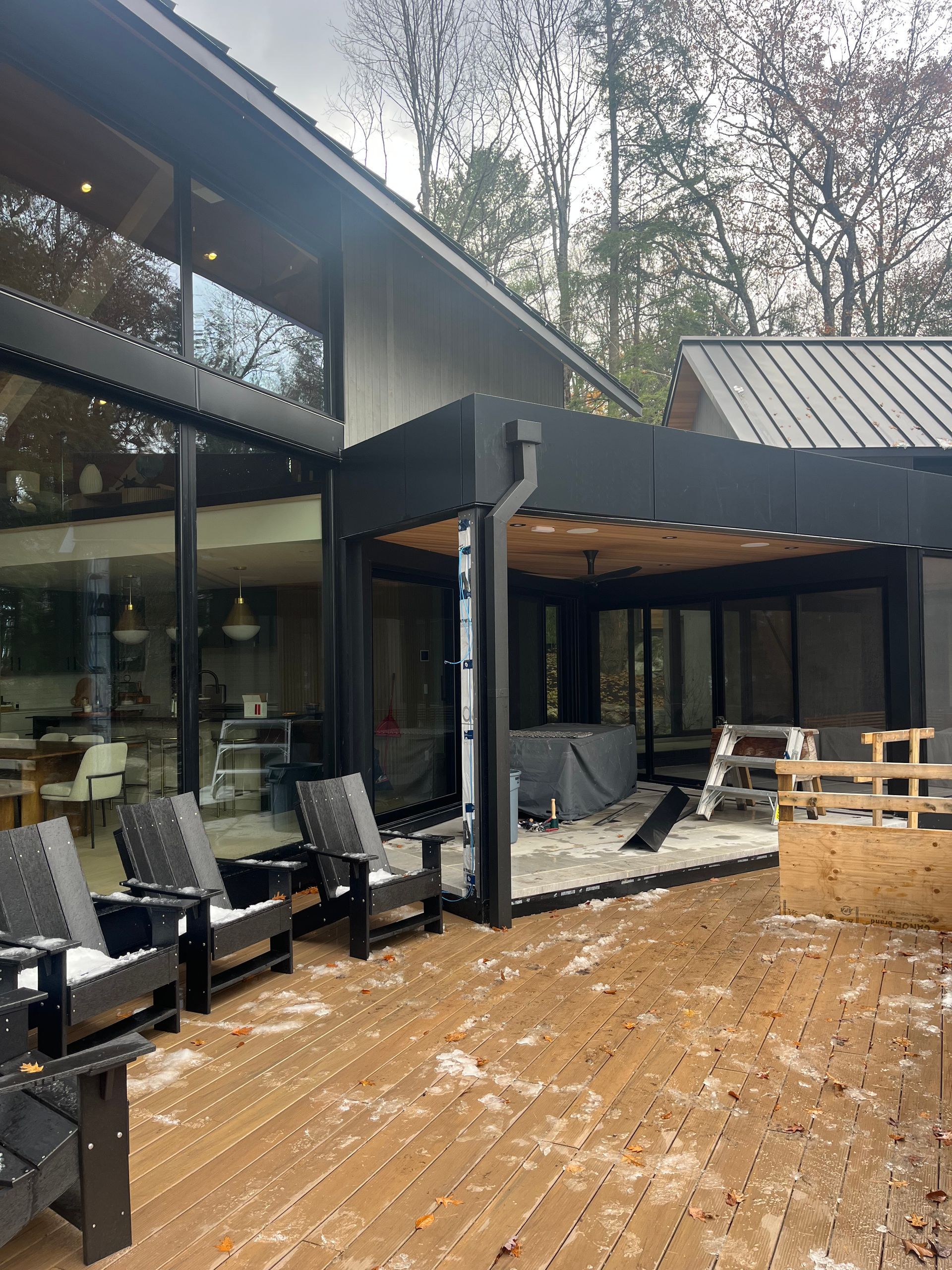 Modern house deck with black lounge chairs, glass walls, and snowy ground surrounded by trees