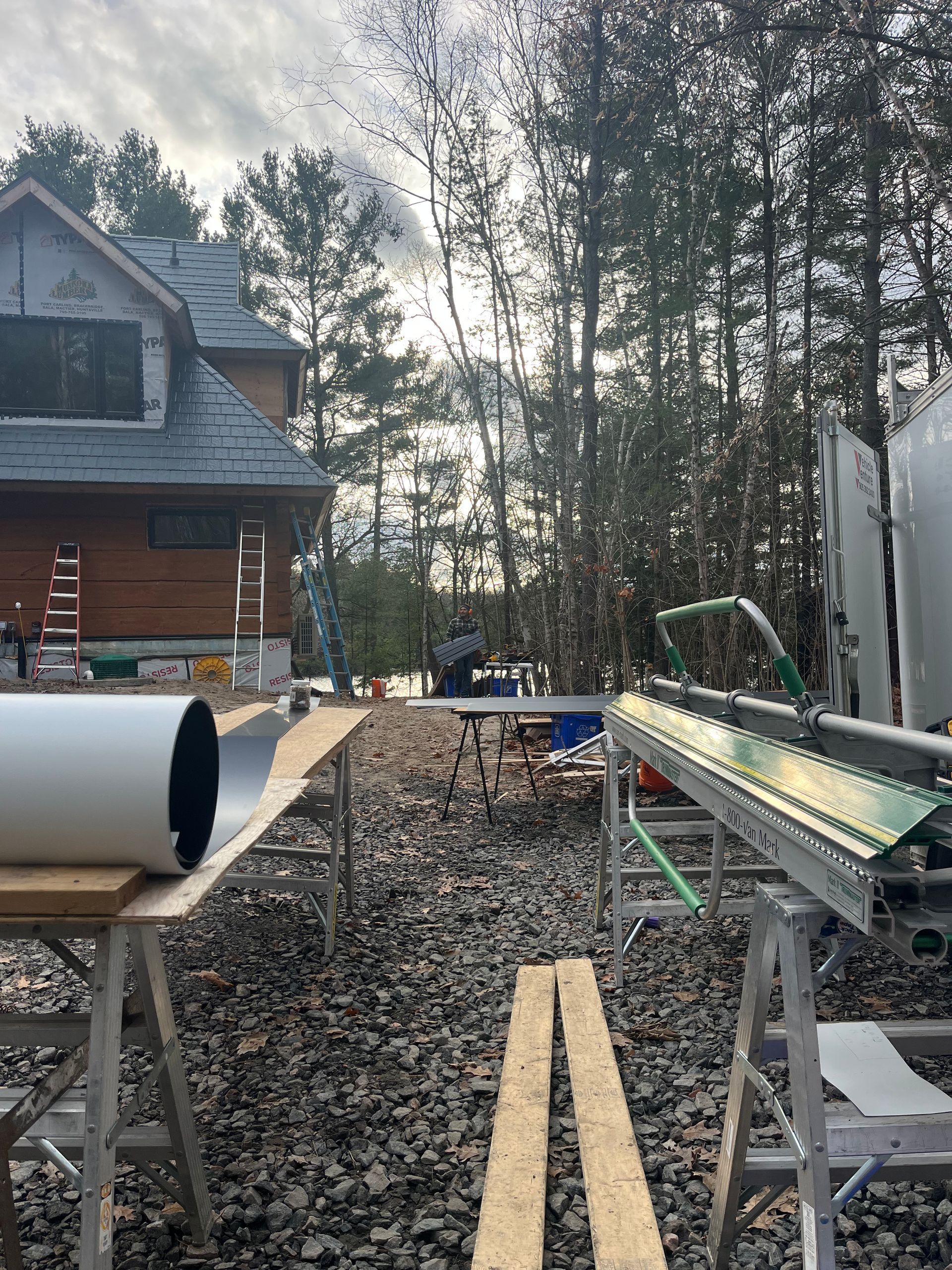 Outdoors by a cabin, a metal rail and pipe sit on stands beside a gravel path and tall trees.