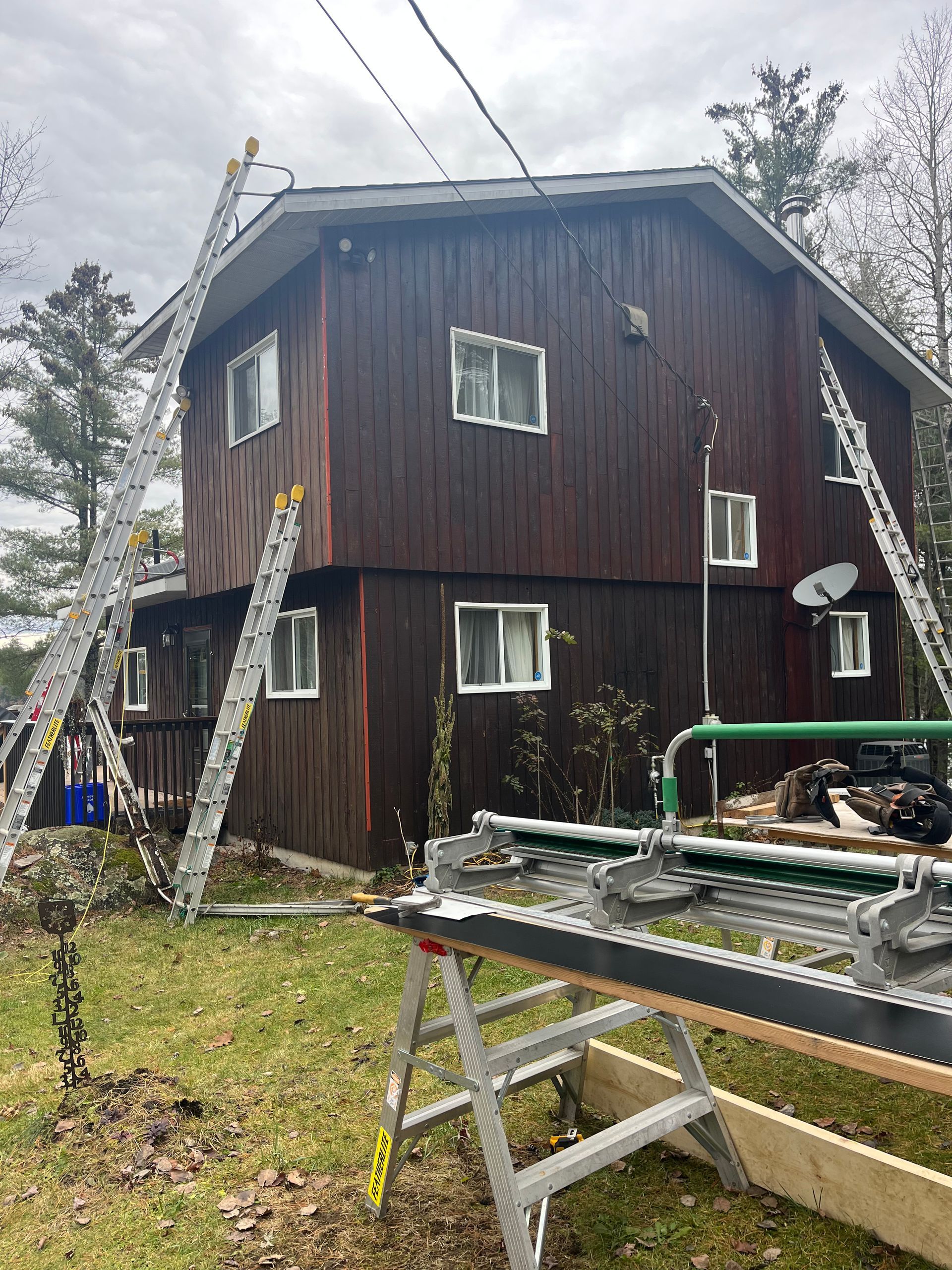 Rustic two-story brown house with white-trimmed windows, ladders, and a work table in the yard under cloudy skies