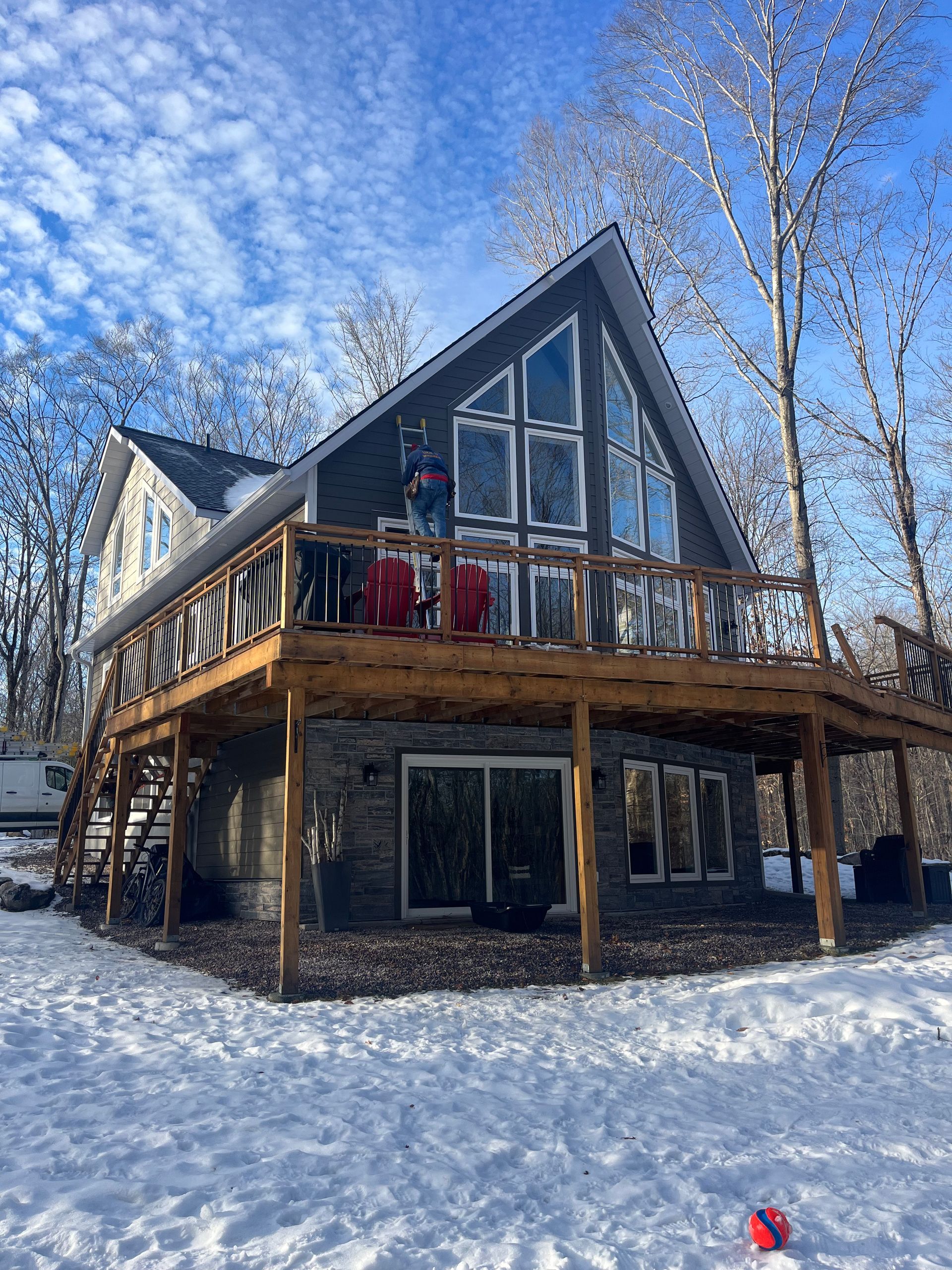 Snowy A-frame cabin with a wraparound deck, tall windows, and red chairs under a bright blue sky