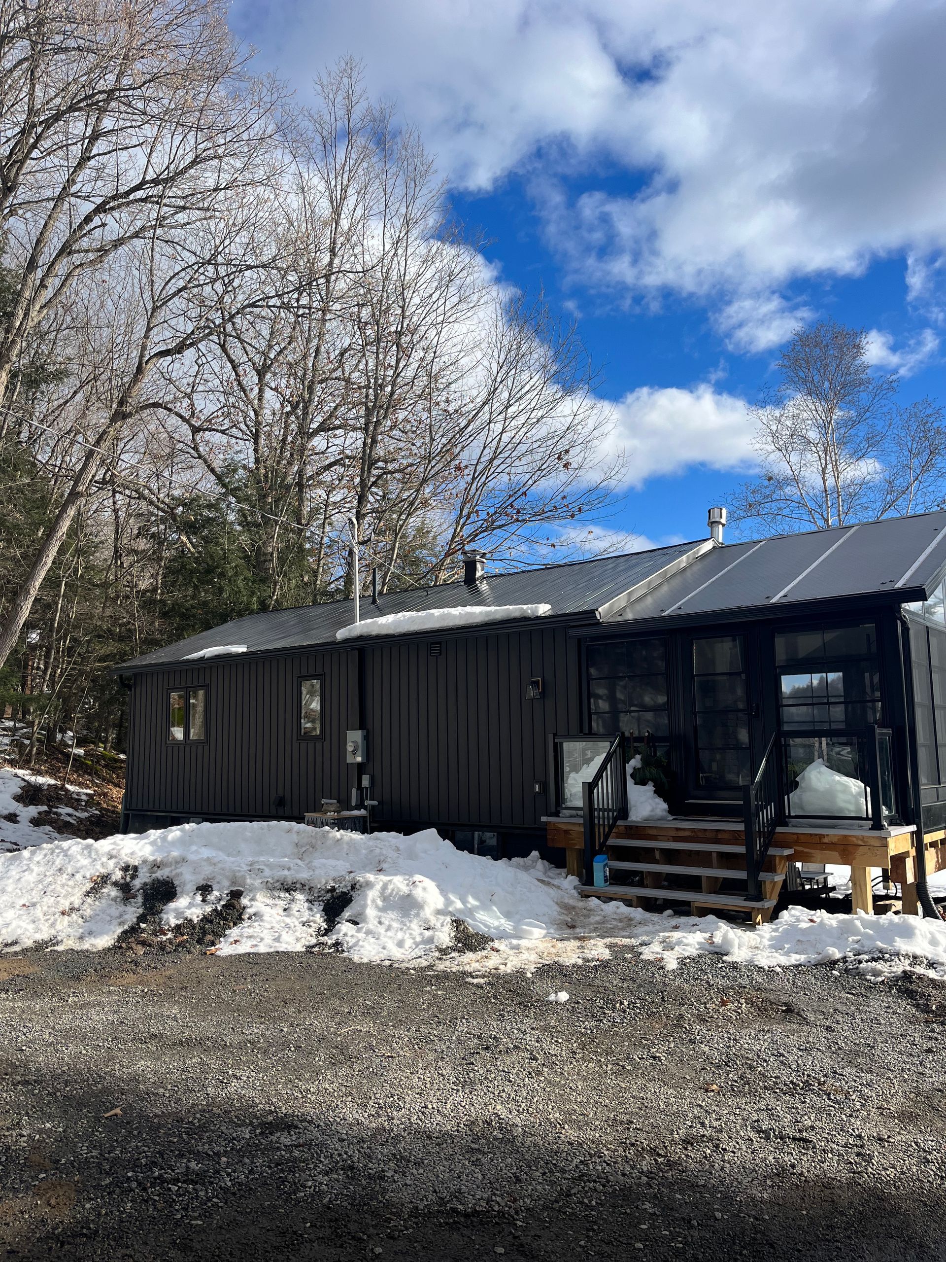 Snowy cabin with dark siding and porch under blue sky, surrounded by leafless trees