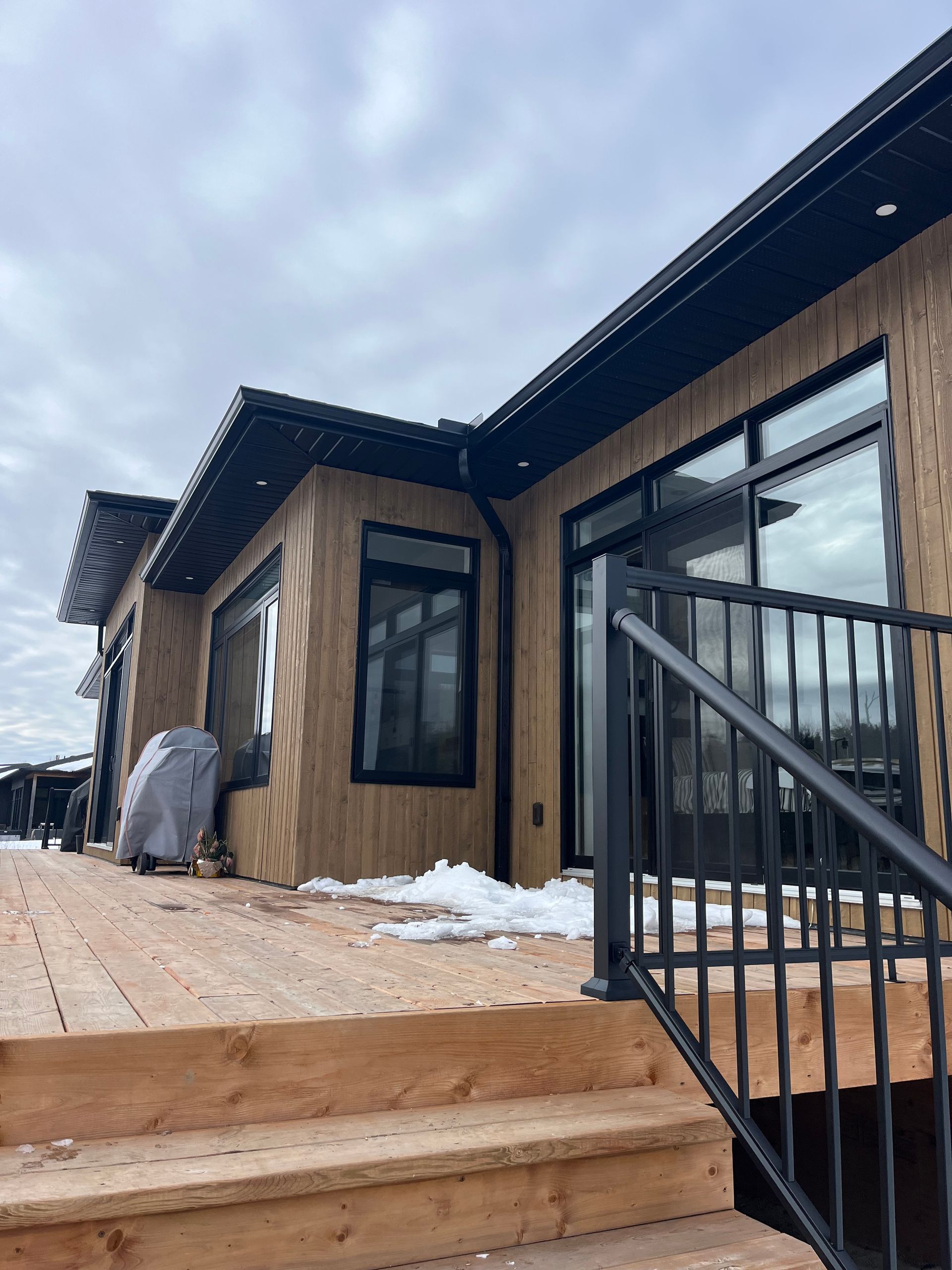 Modern house exterior with wood siding, large windows, and black railing beside a snowy walkway