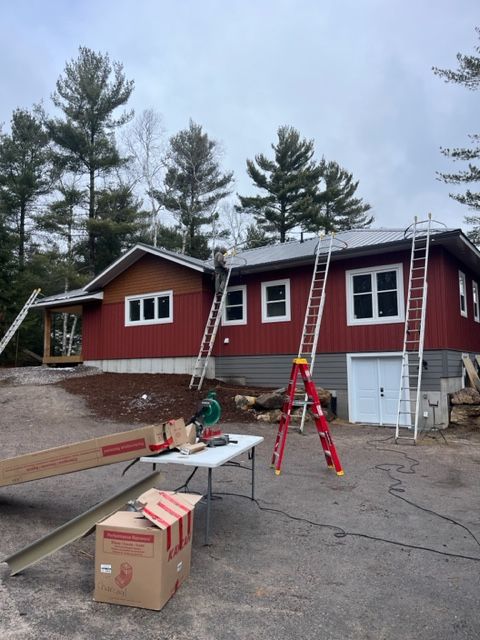 Red cabin under construction with ladders, sawhorse, and boxes in front, pine trees behind.