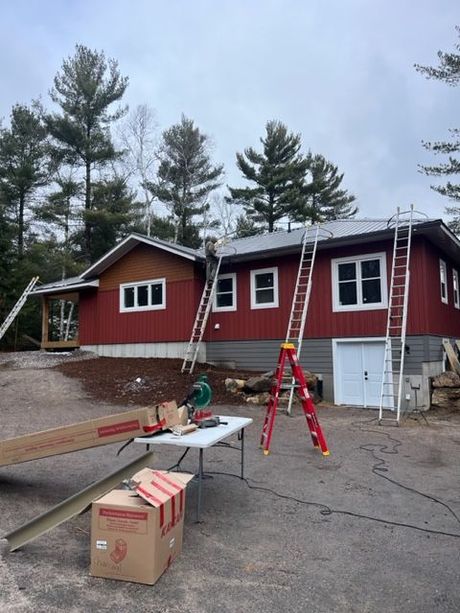Red cabin under construction with ladders, sawhorse, and boxes in front, pine trees behind.