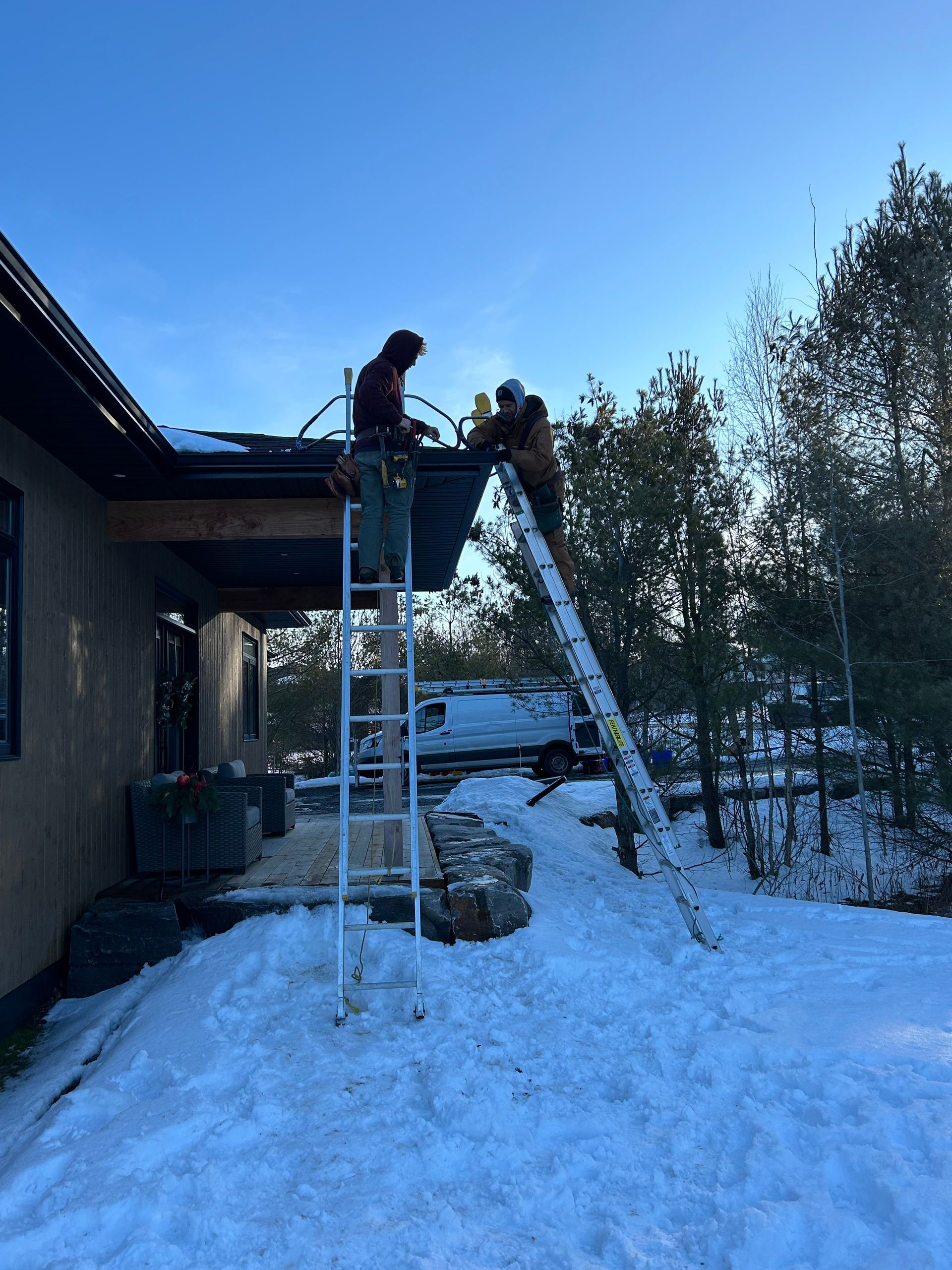 Two people on ladders working on a snowy roof beside a house at dusk