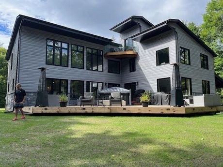 Modern gray two-story house with a wooden deck and lawn, with a person standing at left.