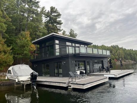 Modern dark-gray lakeside house with wraparound deck and boat docked beside it, surrounded by trees