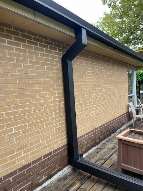 Black downspout and drainpipe on a brick house exterior beside a patio and planter box