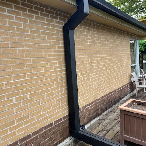 Black downspout on a tan brick house beside a patio with a brown planter box.