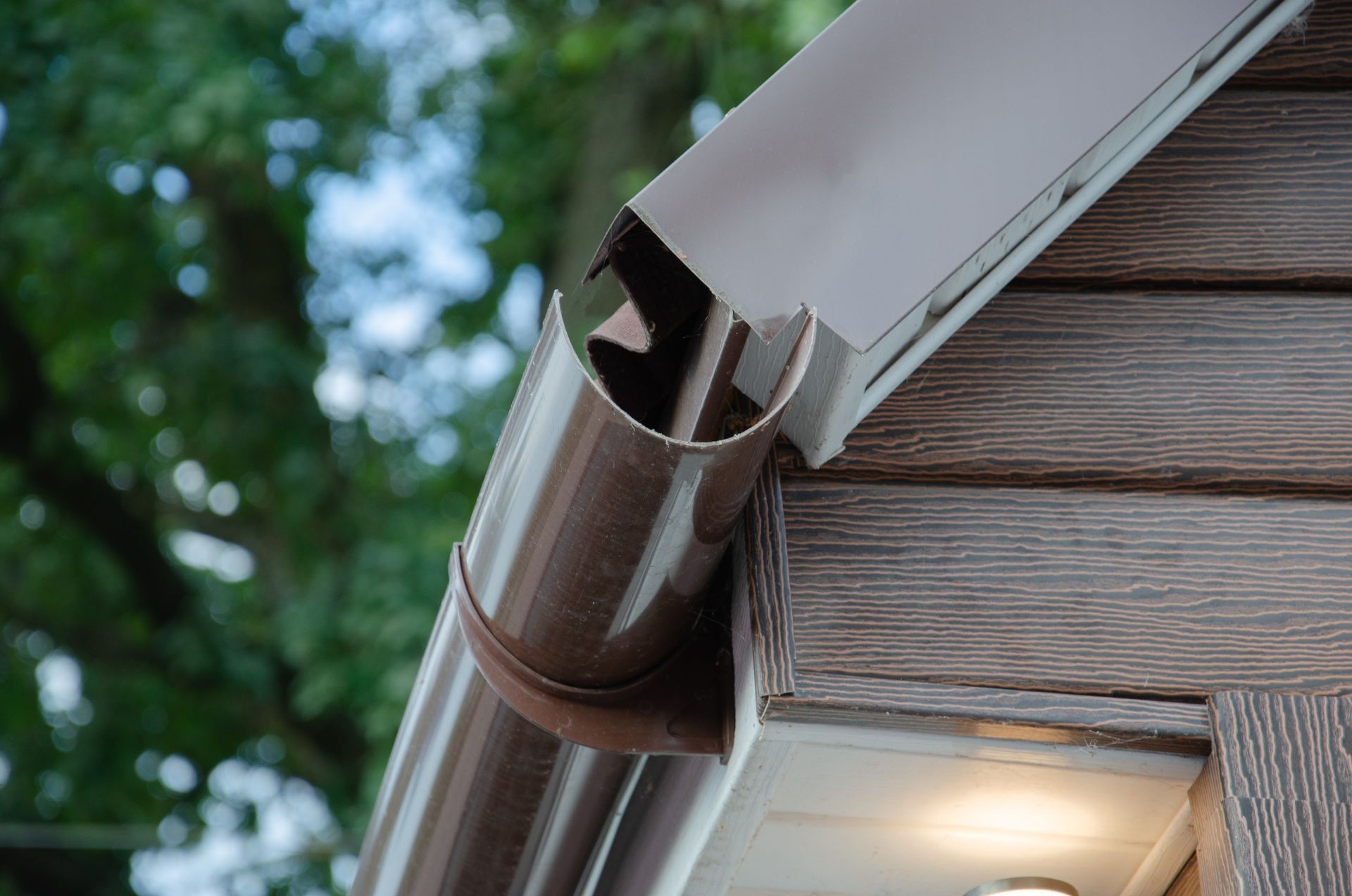 Brown gutter and downspout on a house roof edge against a leafy background
