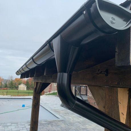 Black gutter and downspout attached to a wooden roof edge above a paved yard
