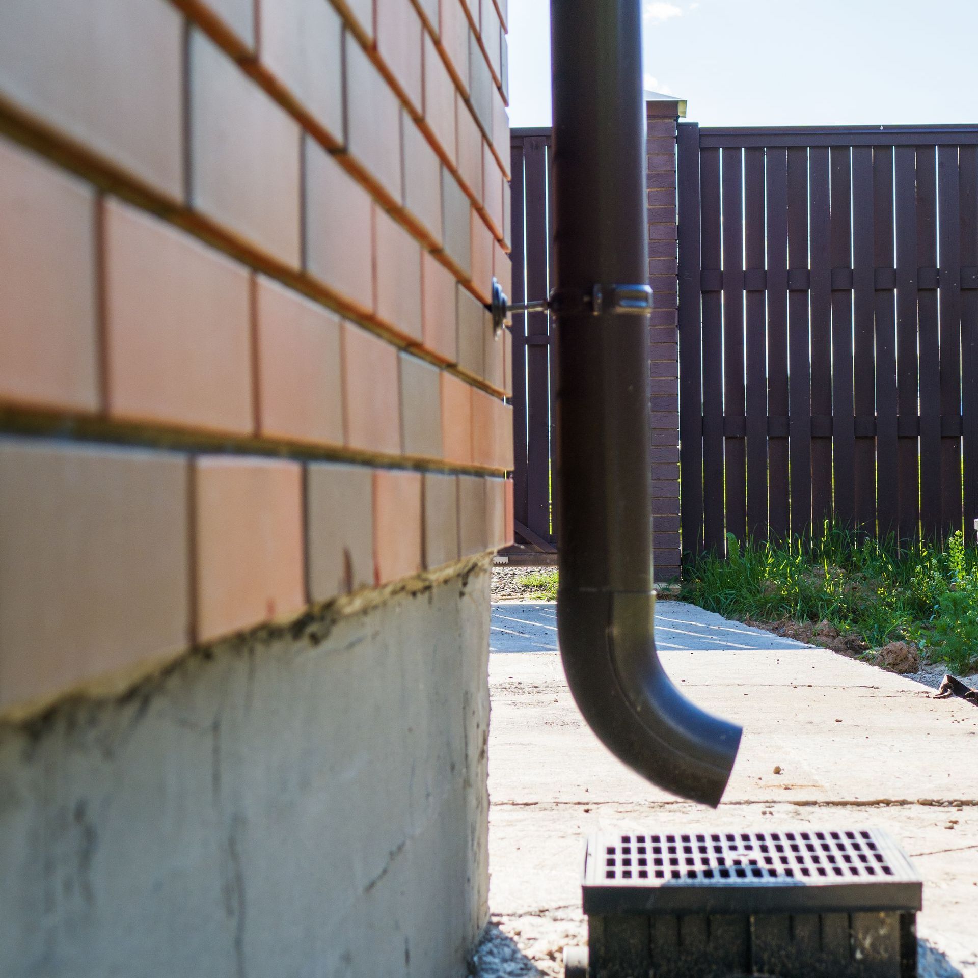 Side yard with brick wall, downspout, concrete path, and drain grate beside a wooden fence.
