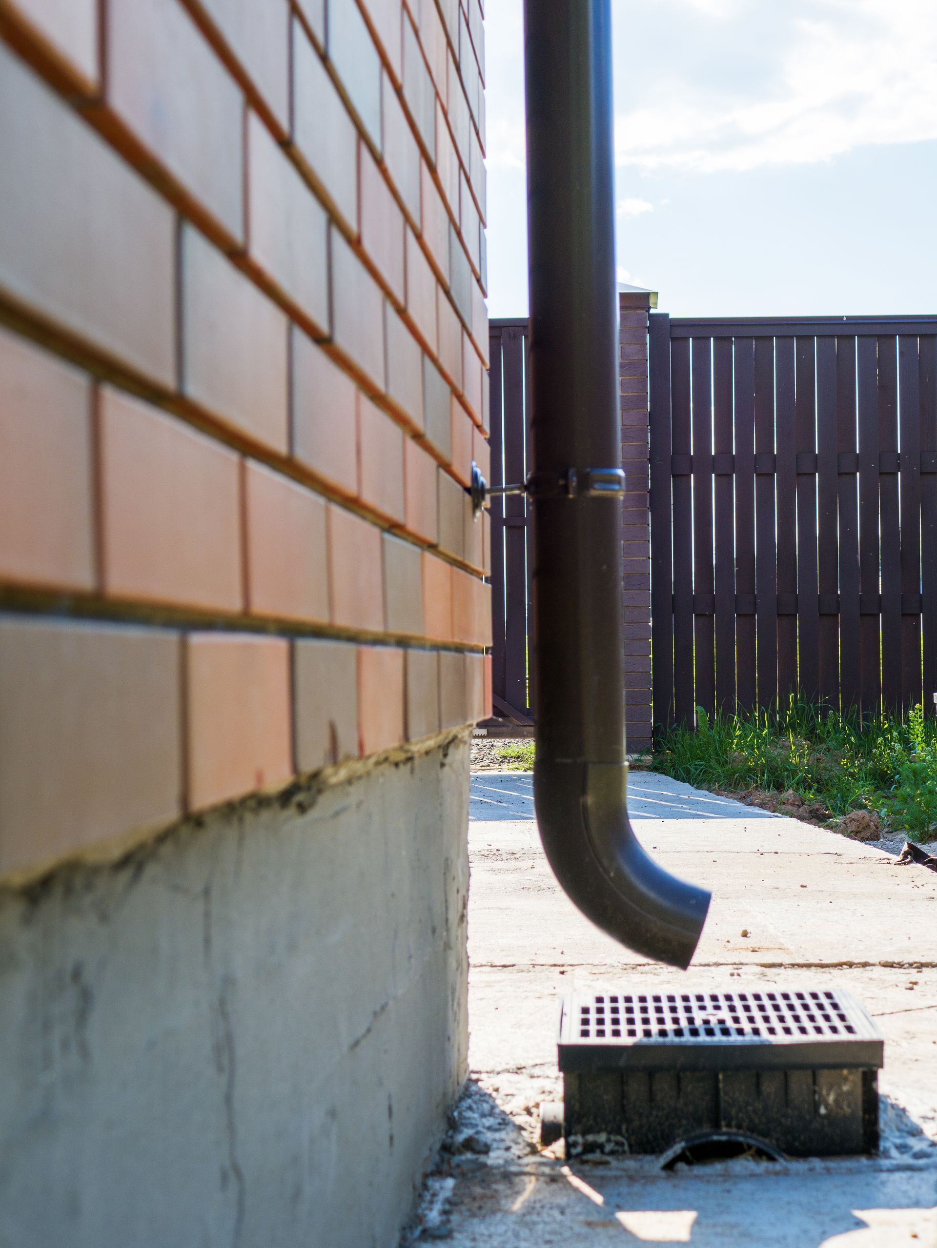 Brick wall, downspout, and drain grate in a narrow outdoor alley beside a wooden fence.