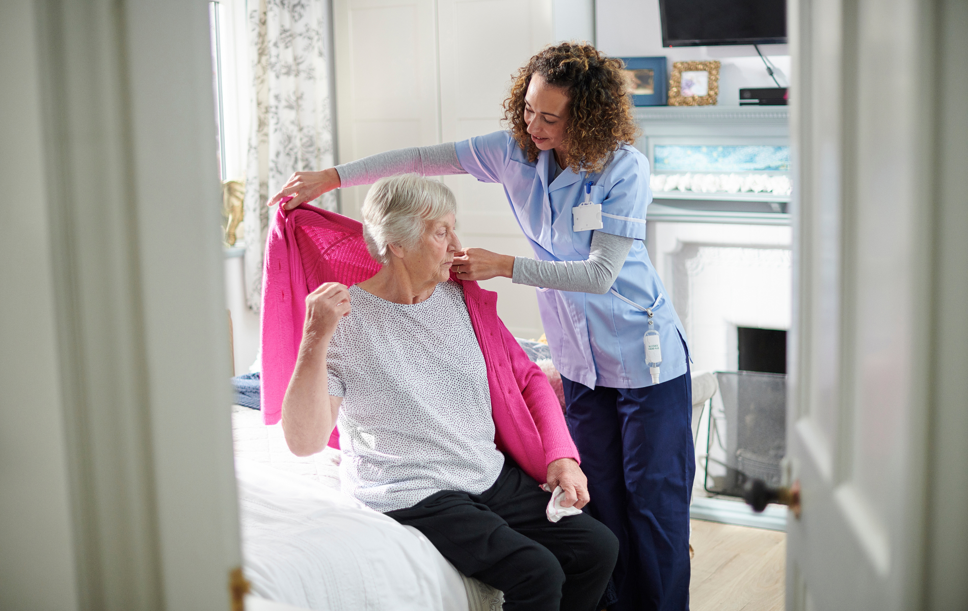 Caregiver helping an elderly woman put on a pink cardigan in a bright bedroom