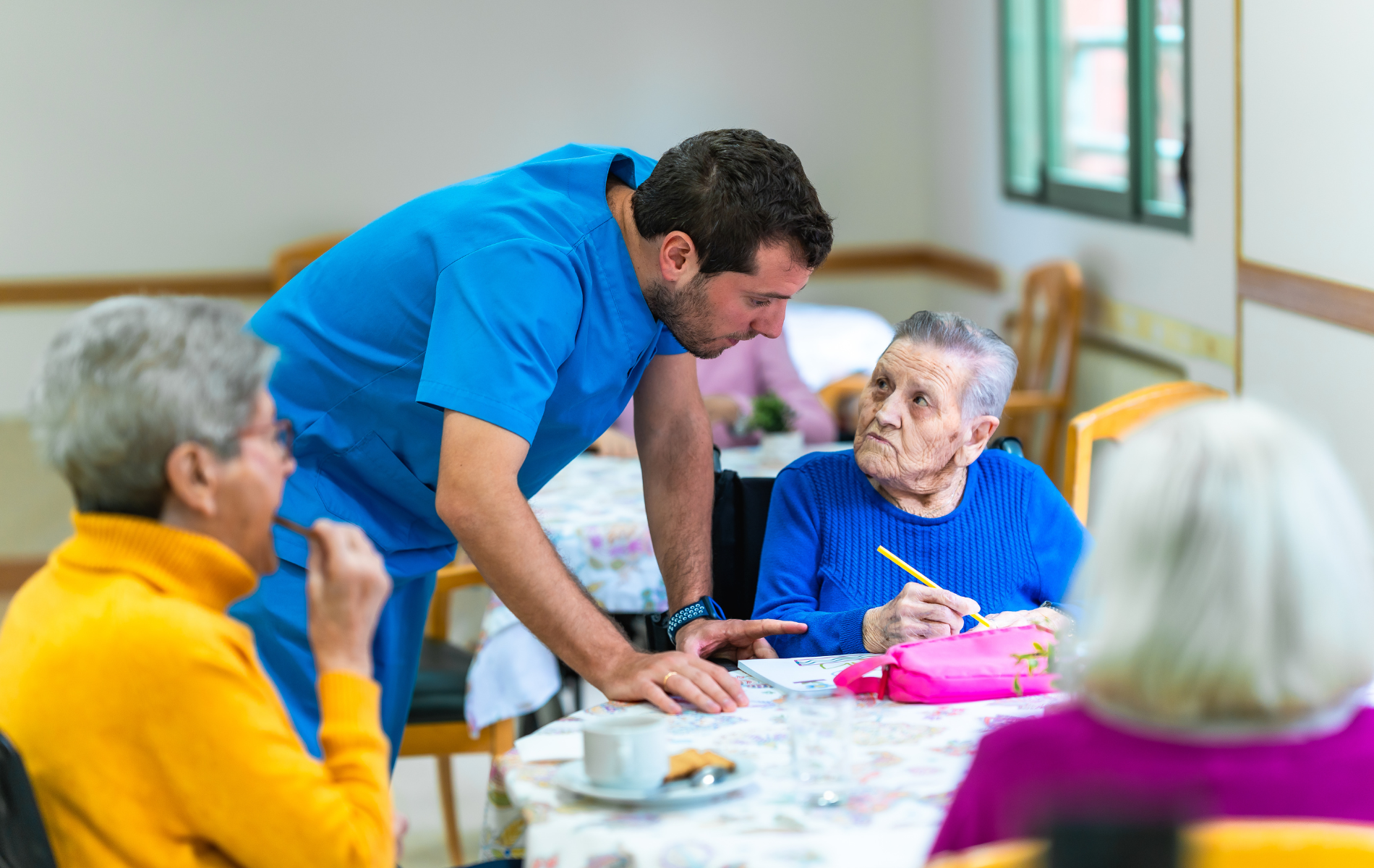 Caregiver serving food to residents at a table in a bright dining room