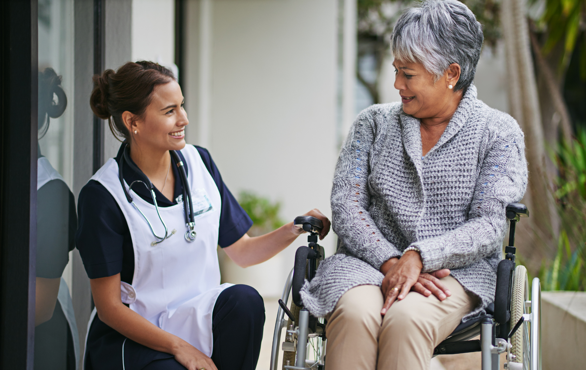 Caregiver in white uniform talks with smiling patient in wheelchair outside a building