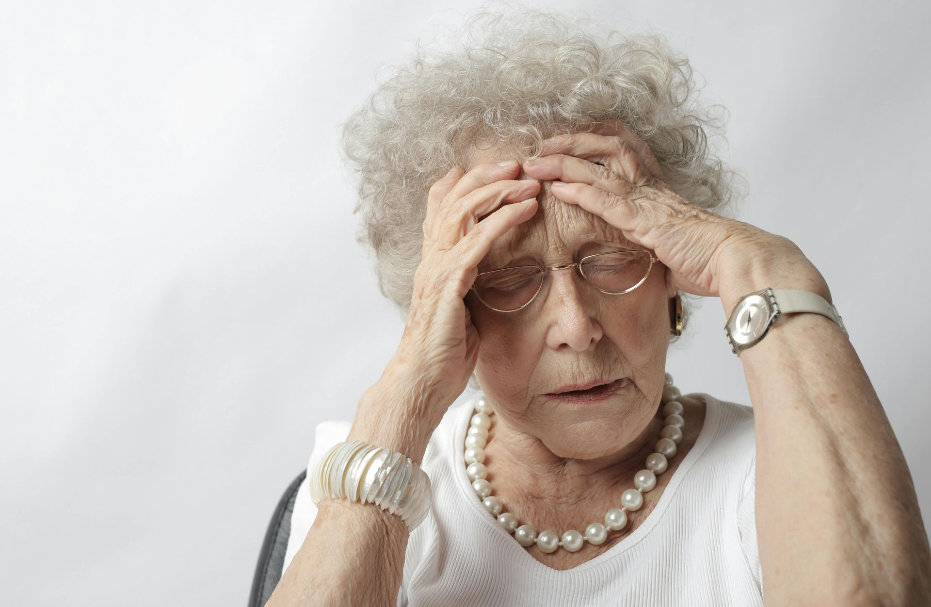 Older woman holding her head with a concerned expression, wearing glasses and a white shirt against a light background