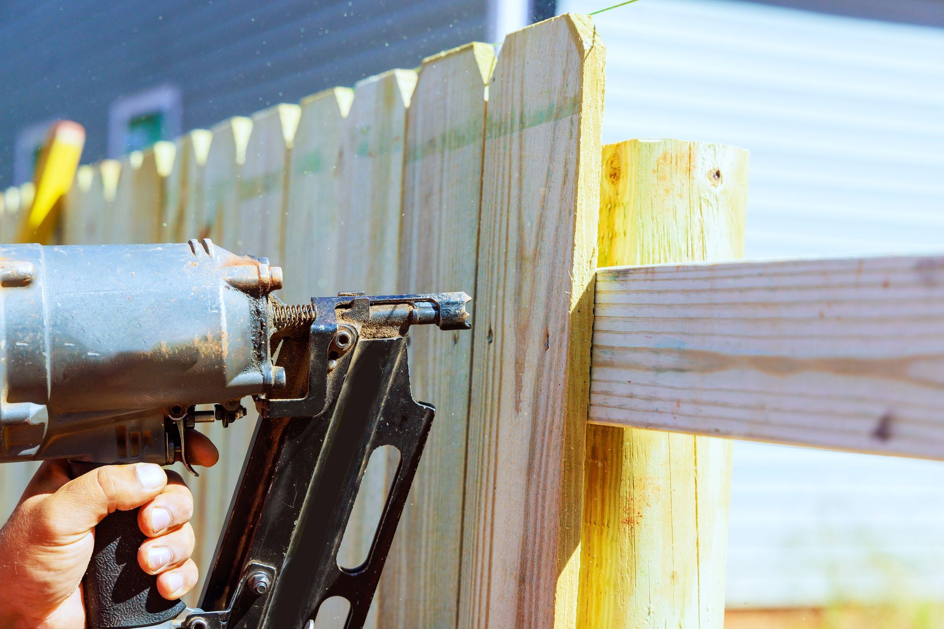 A man uses an air nail gun to construct sections of a wooden fence around a yard.