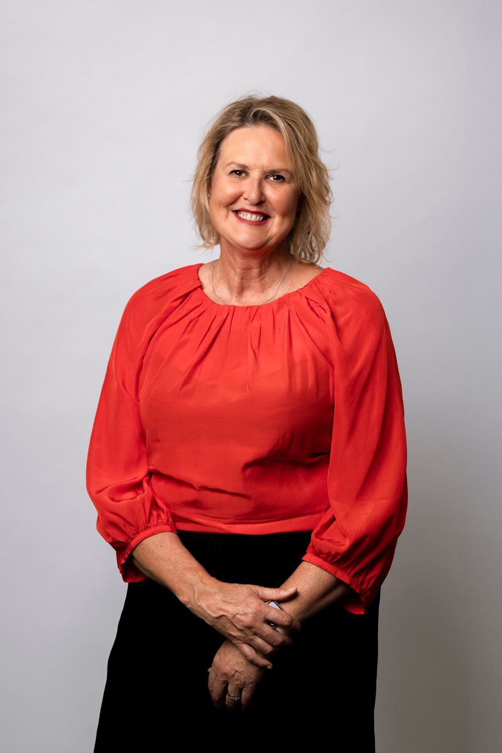 Woman in a red blouse, smiling and seated against a light gray background.