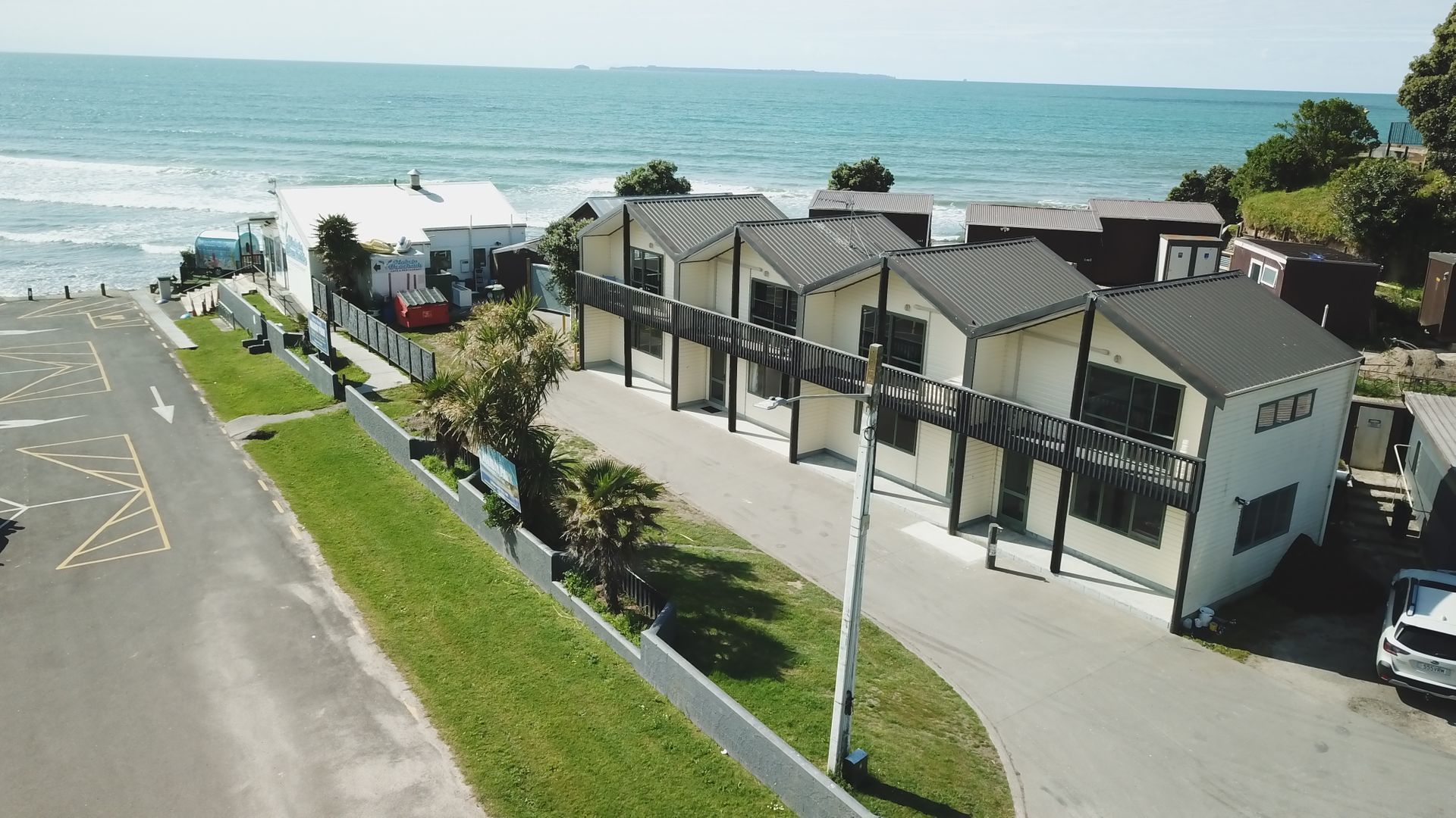 Coastal row of houses beside a road, with a grassy strip and ocean waves in the background.
