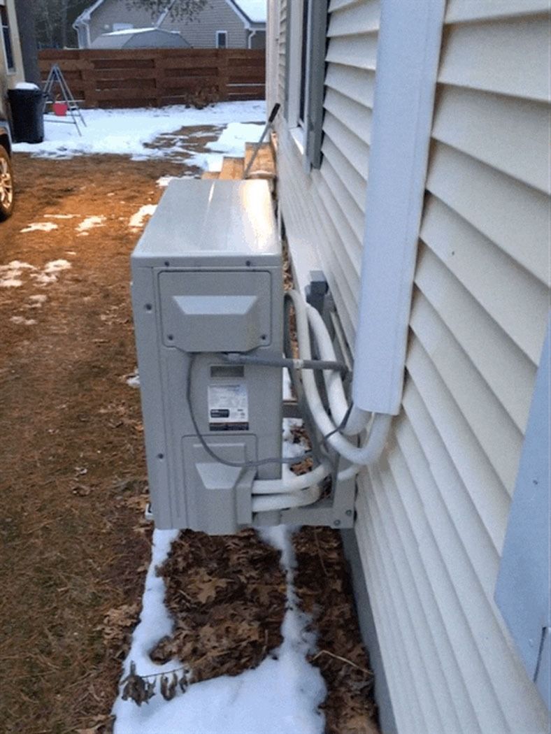 Outdoor HVAC unit beside a house wall with pipes and snow on the ground