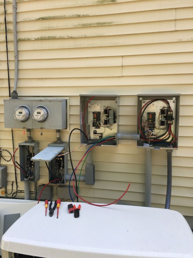 Open electrical panels and gauges mounted on an exterior wall above a white workbench with wires.