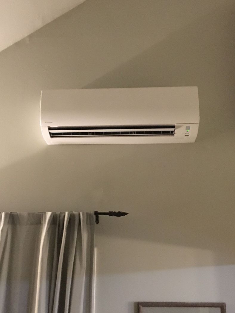 Wall-mounted white air conditioner above a curtained window in a beige room.