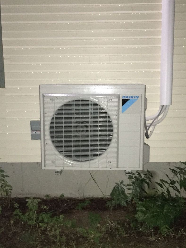 Outdoor air conditioner unit mounted on a house wall beside a white downspout.