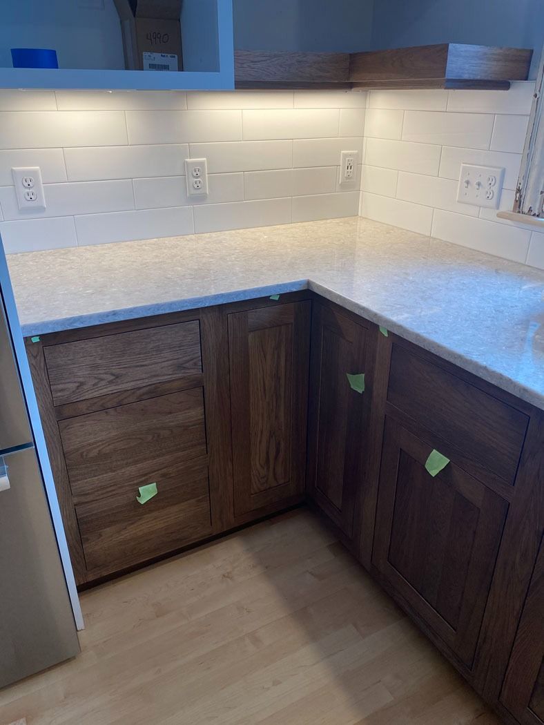 L-shaped kitchen corner with white tile backsplash, light speckled countertop, and dark wood cabinets under construction