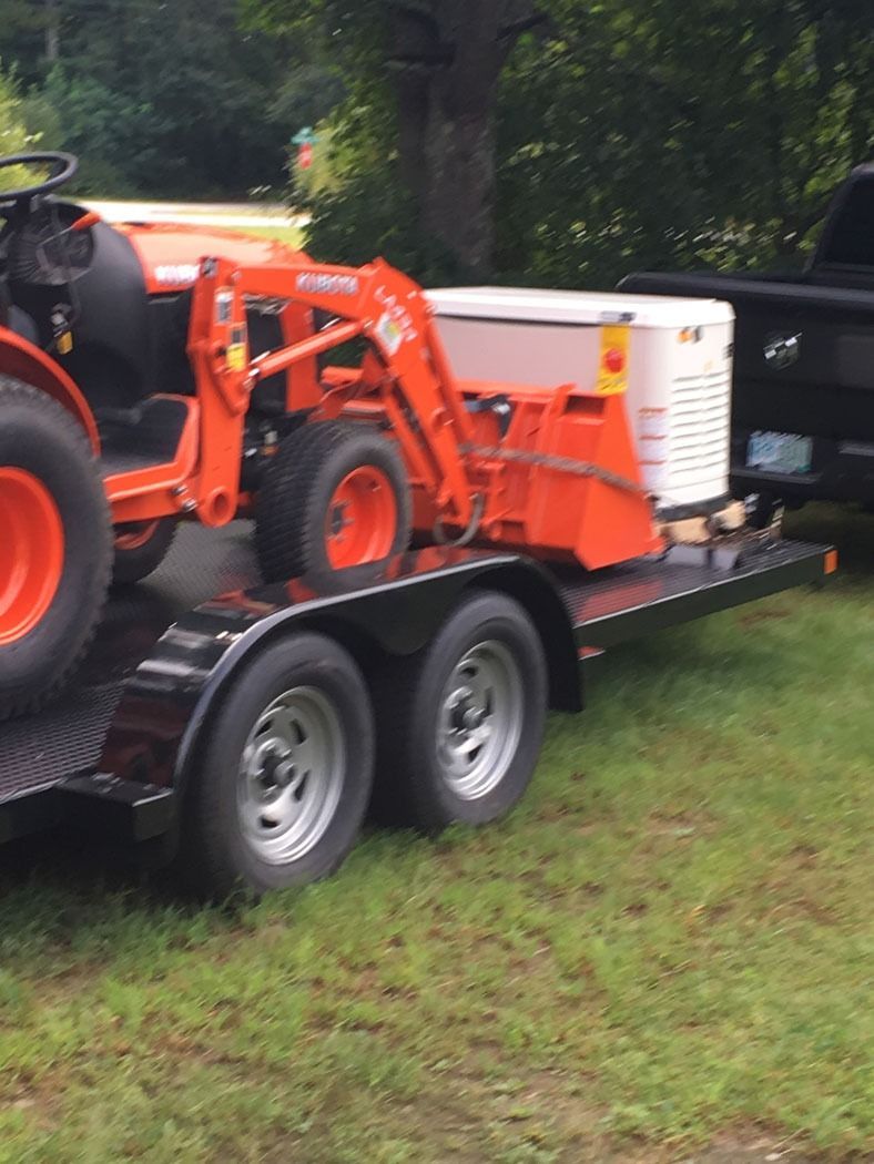 Orange tractor and front loader strapped on a black trailer in a grassy yard.