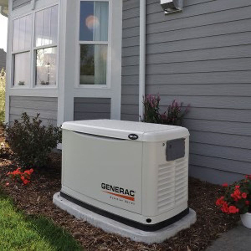 Generac standby generator beside a house, on a concrete pad near gray siding and red flowers