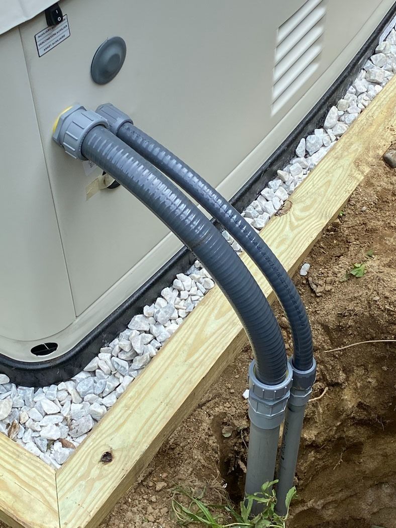 Two gray conduit pipes entering an outdoor utility unit beside a gravel bed and wooden border.