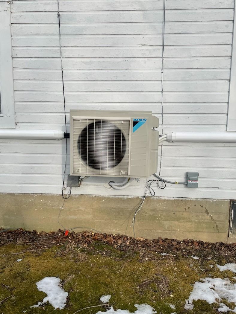 Outdoor air conditioner unit mounted on a white wall above a concrete foundation, with snow on the ground