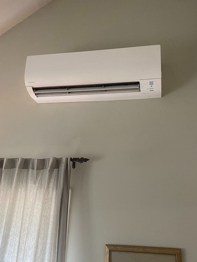 Wall-mounted white air conditioner above a window with sheer curtains in a light-colored room