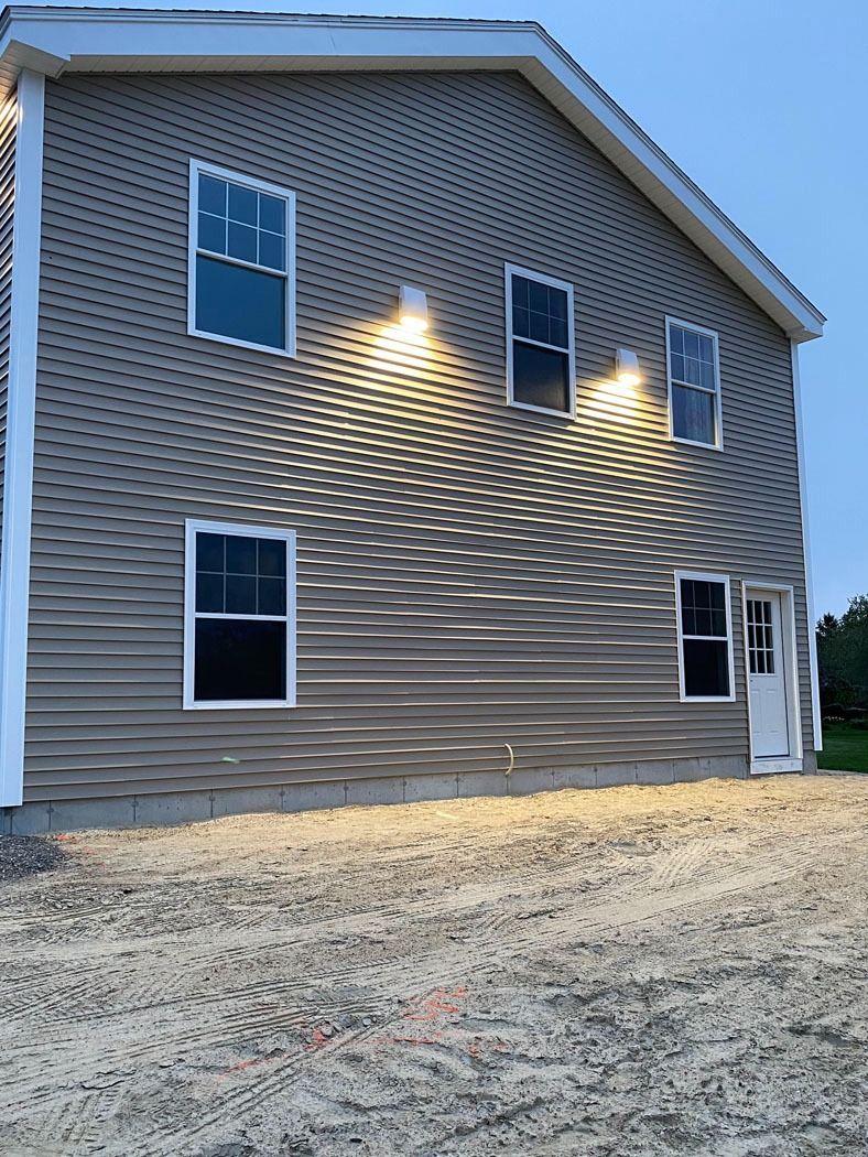 Two-story tan house with white trim, lit by exterior lights, seen from the side on gravel.