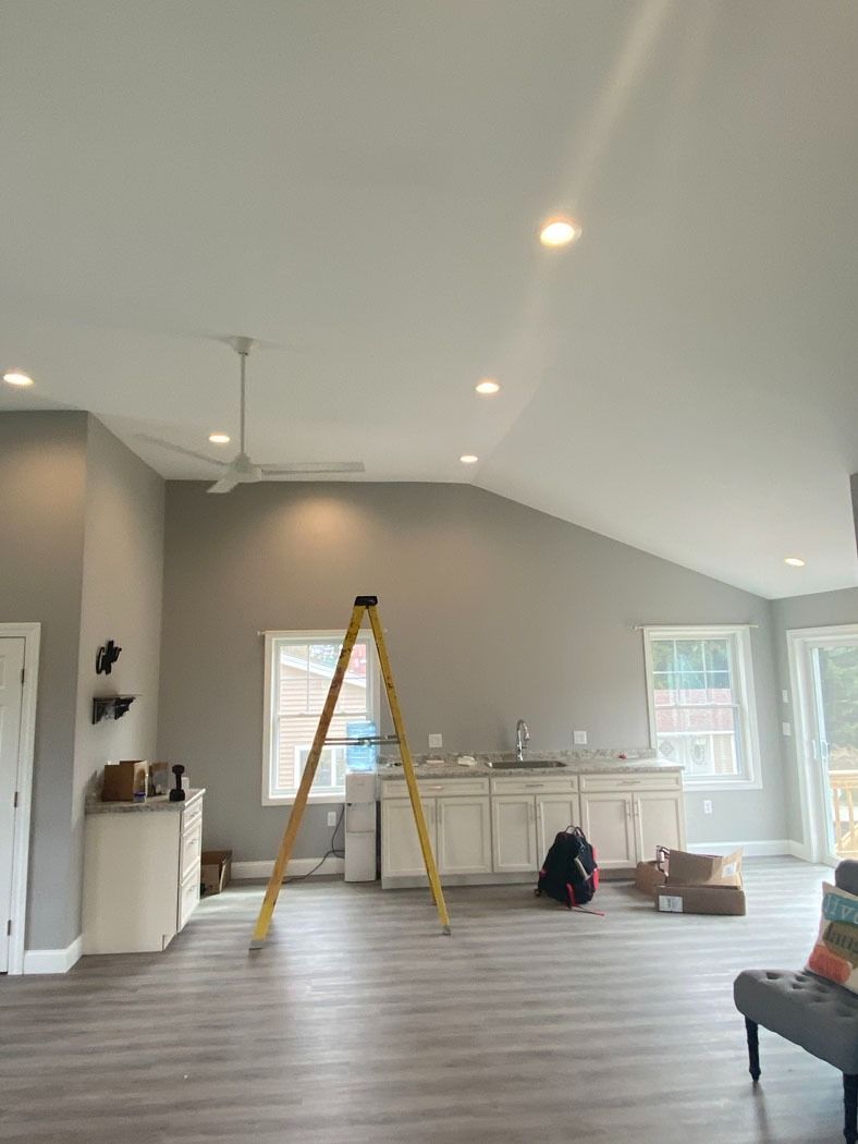 Bright living room with sloped white ceiling, gray walls, ladder, and cabinets under windows