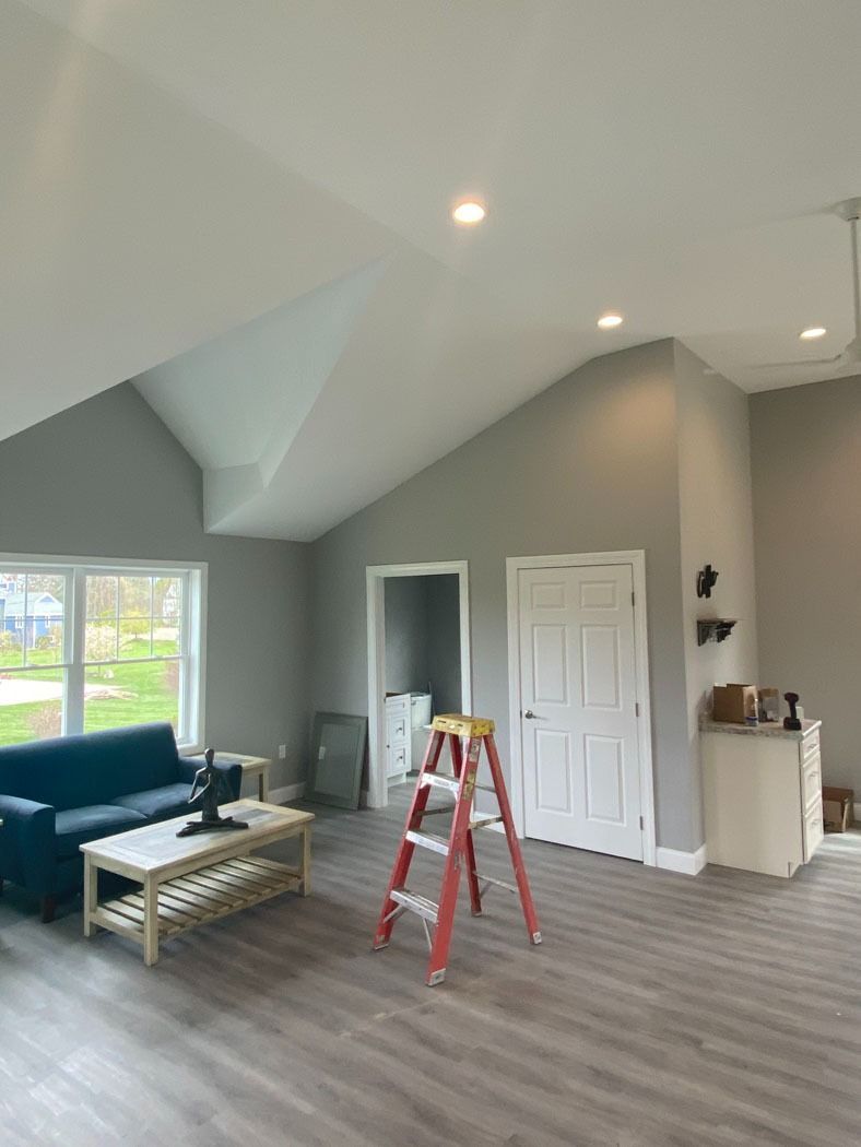 Spacious gray living room with vaulted ceiling, blue sofa, ladder, and open doorway.