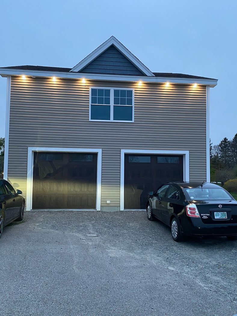 Two-car garage with lit exterior at dusk, gravel driveway, and parked cars in front of a gray house