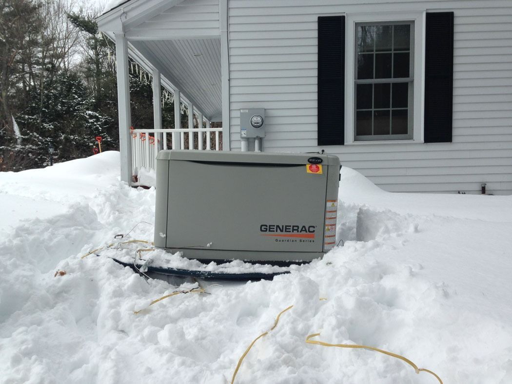 Gray generator sitting in snow beside a white house and porch with black shutters.