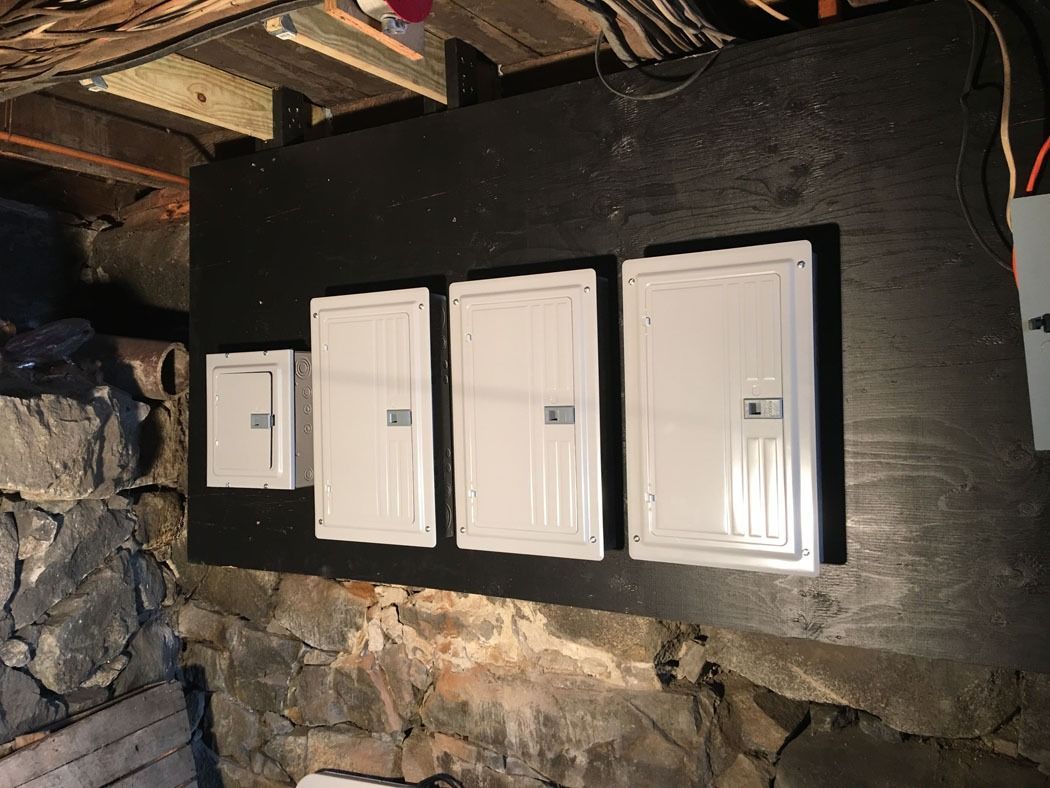 Row of electrical breaker panels mounted on a black board in a basement wall, with exposed ceiling above