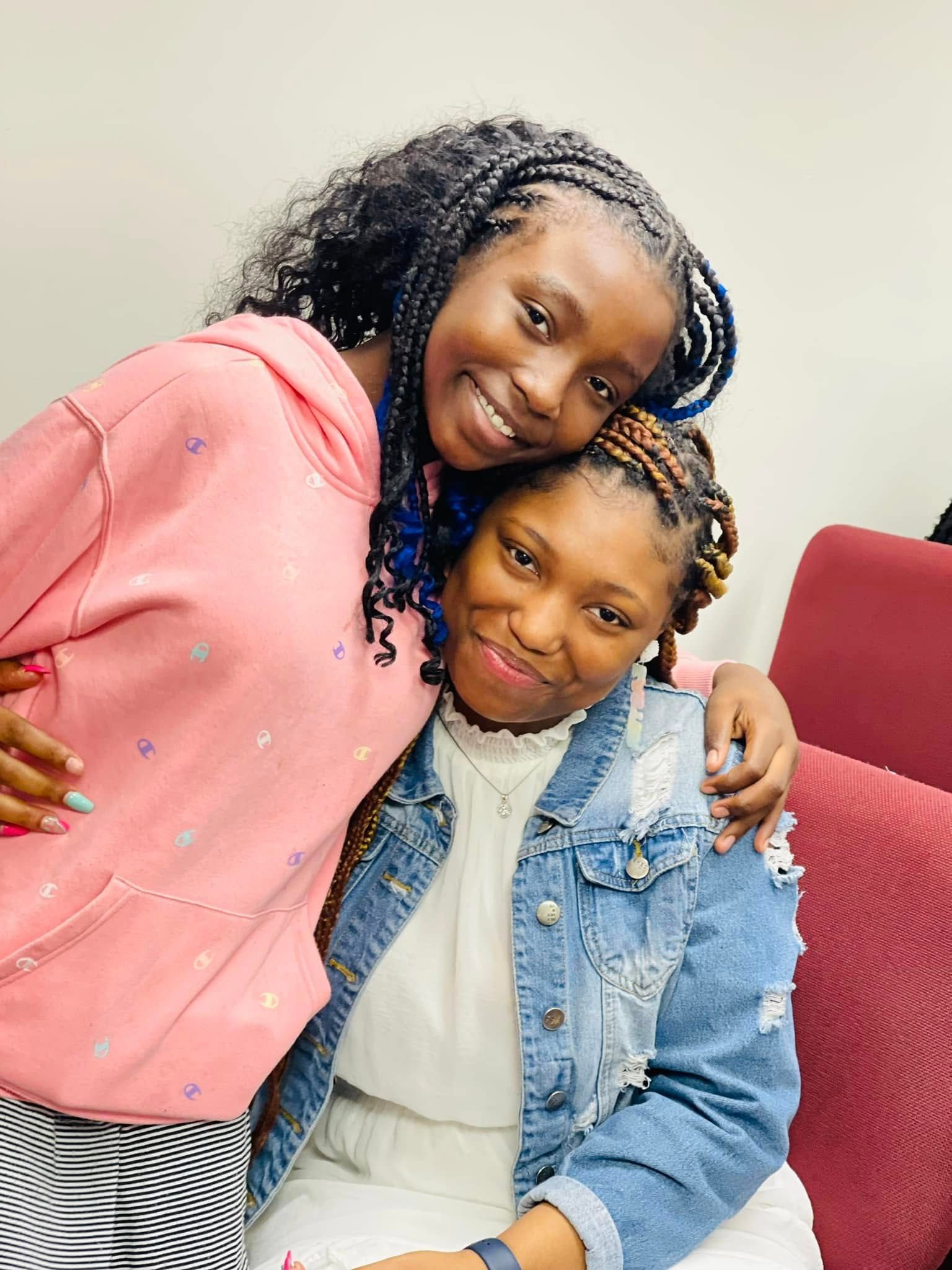 Two people smile as they lean their heads together for a portrait. One wears a pink hoodie and the other a denim jacket.