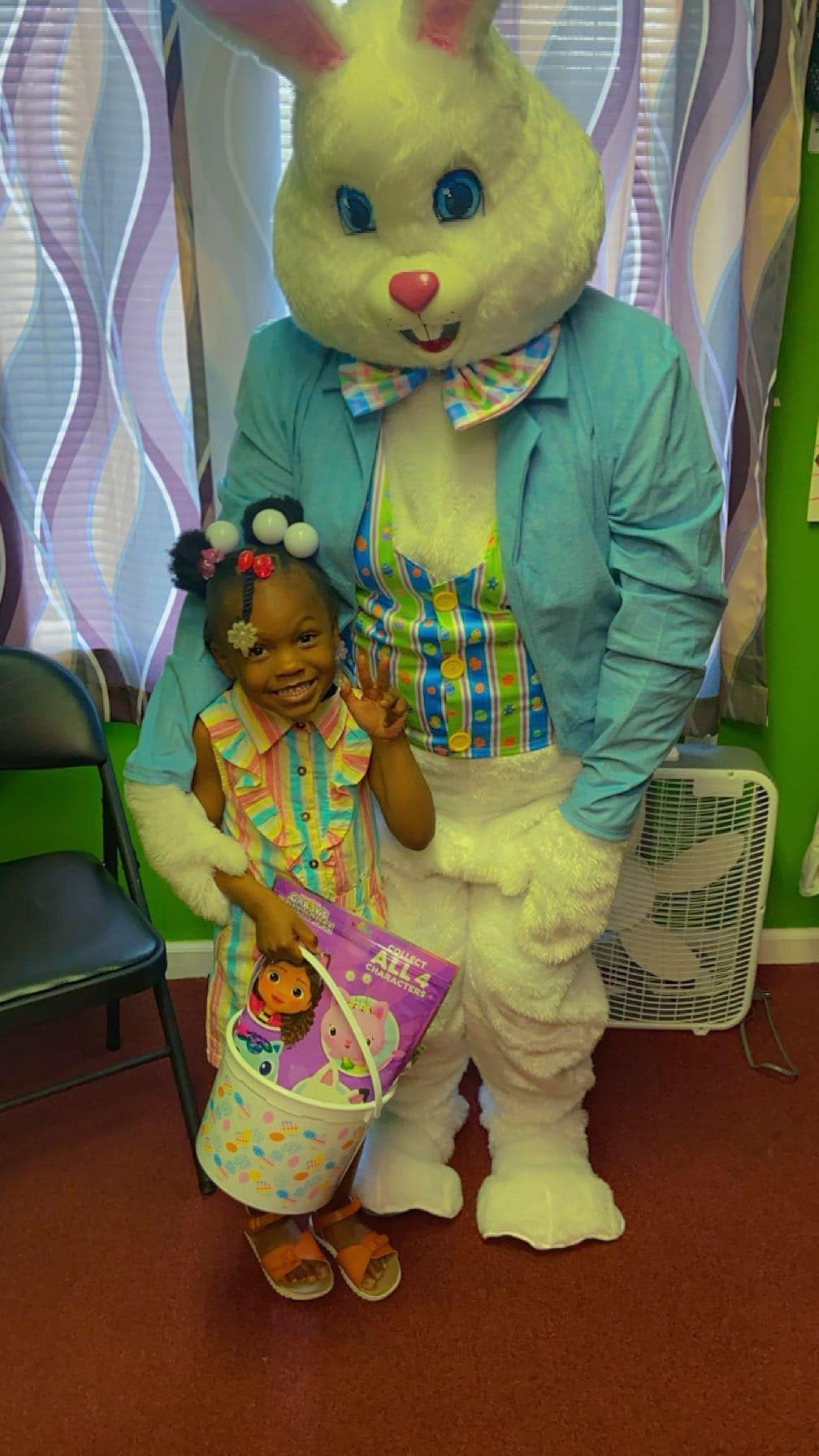 A smiling child stands next to a costumed Easter Bunny, holding an Easter basket with a toy, indoors against curtains.
