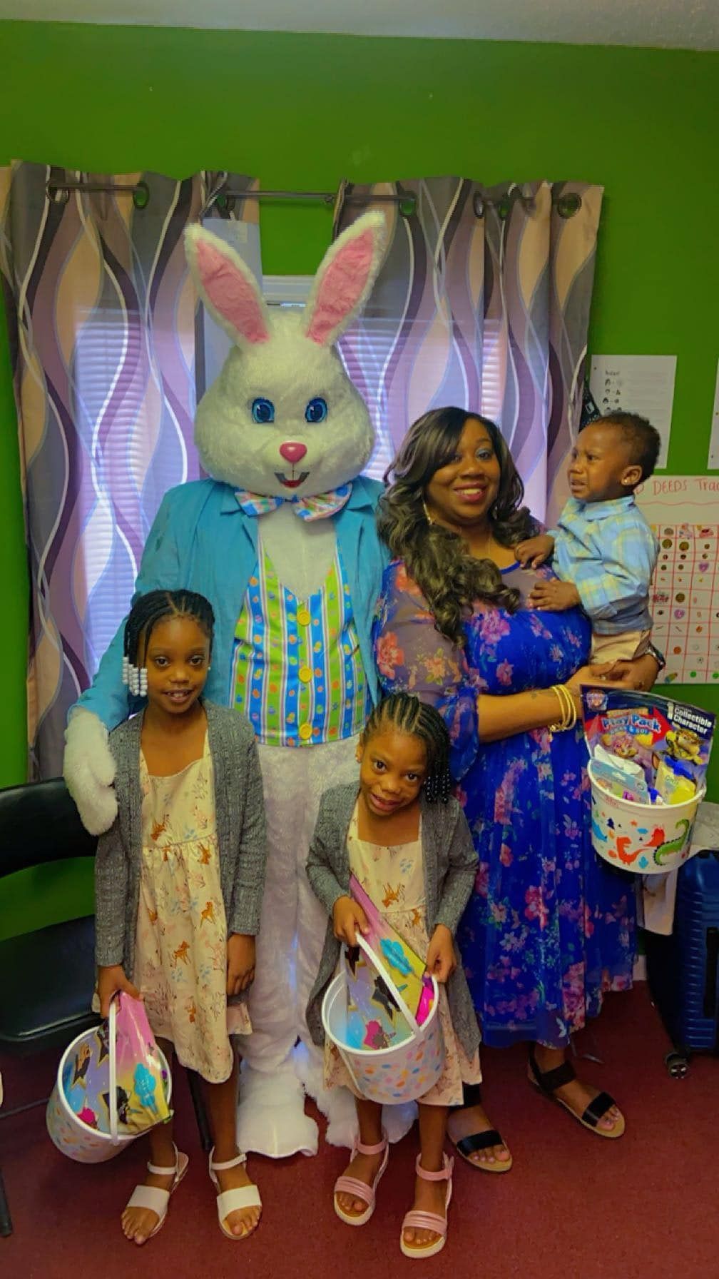 A family poses with an Easter Bunny mascot inside against a window with patterned curtains, holding Easter baskets.