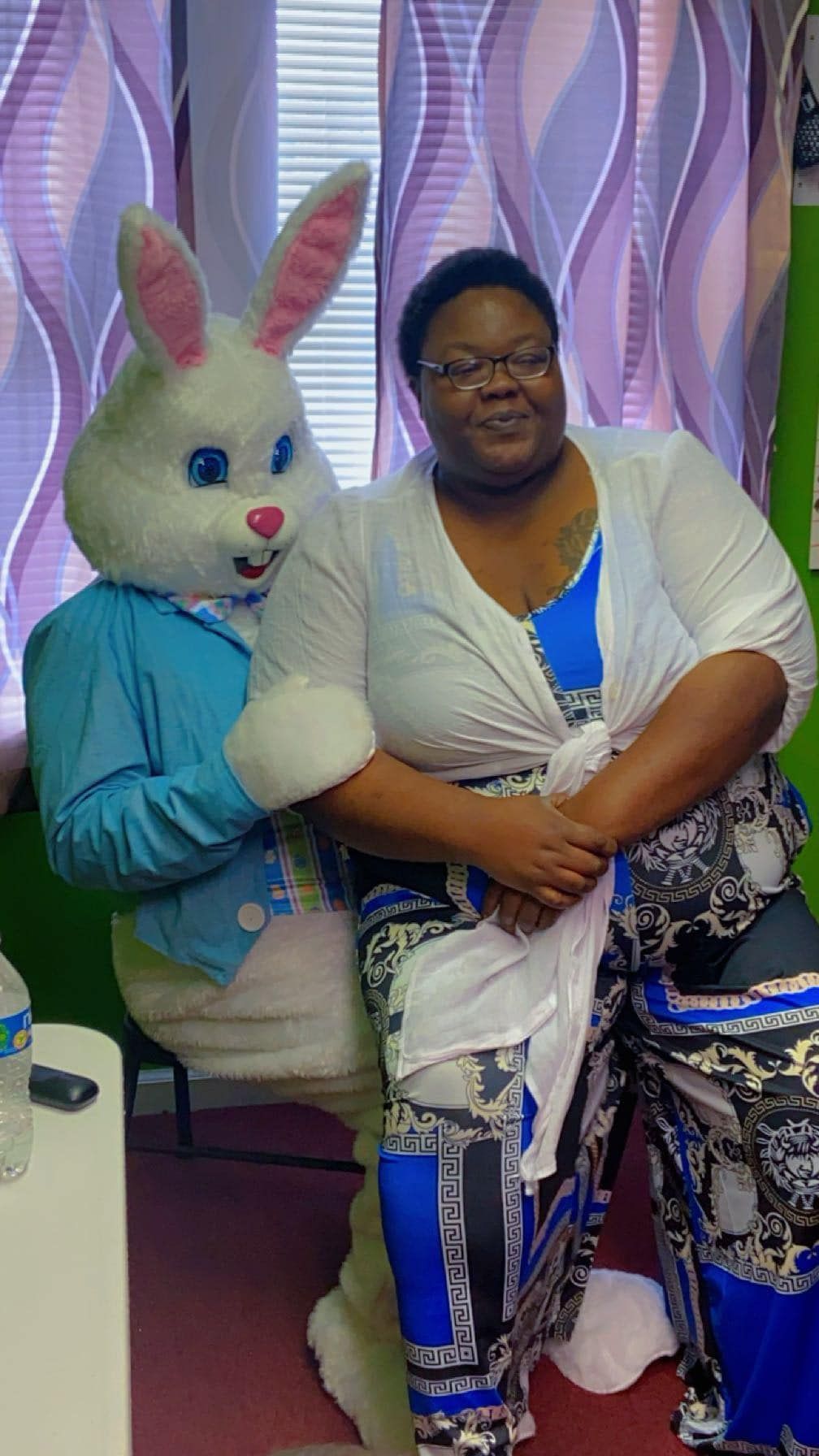 A person with glasses sits smiling next to a person in a white rabbit costume wearing a blue jacket in an indoor room.