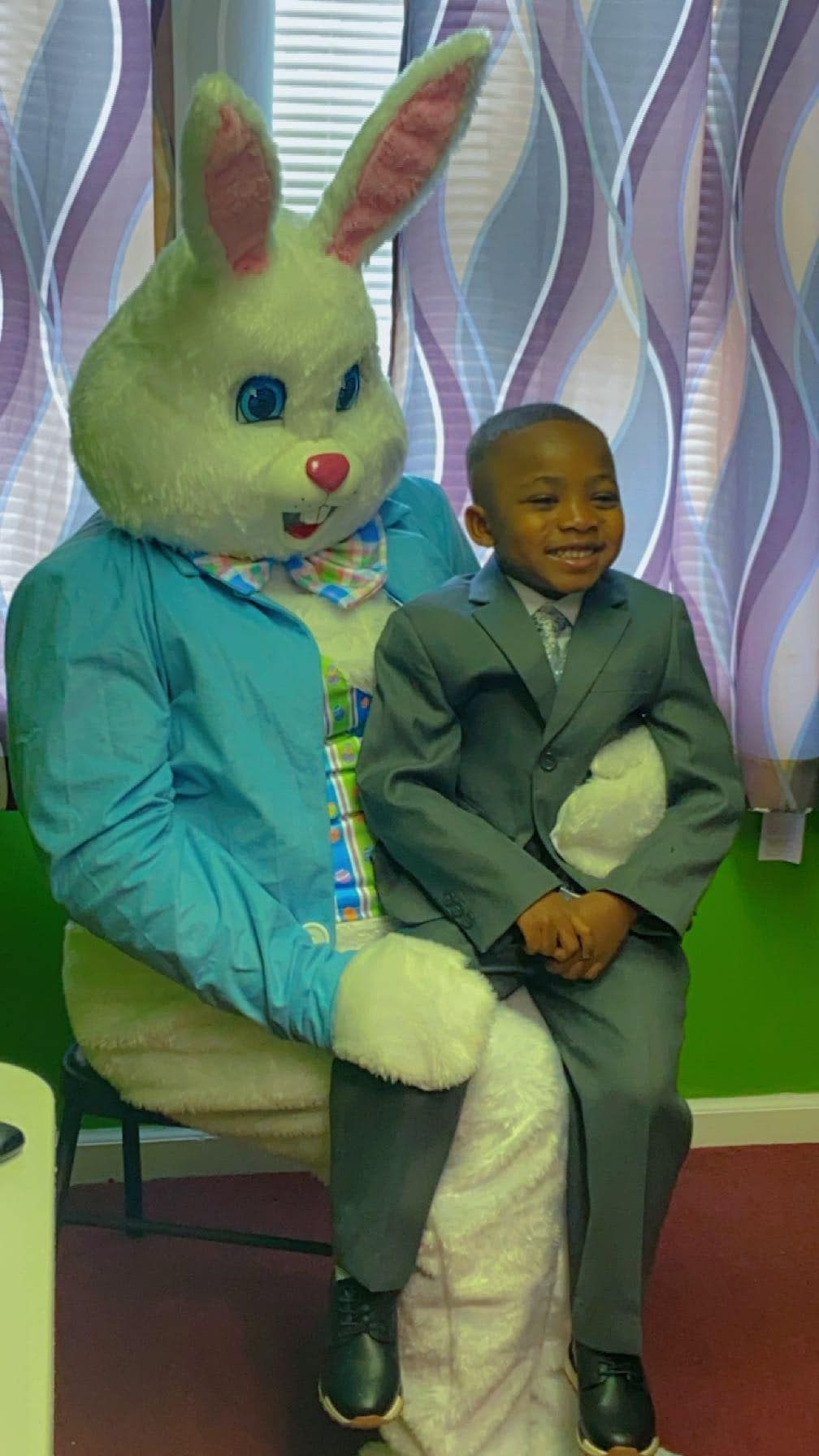 A young child in a suit smiles while sitting on the lap of a person in a full-body Easter bunny costume.
