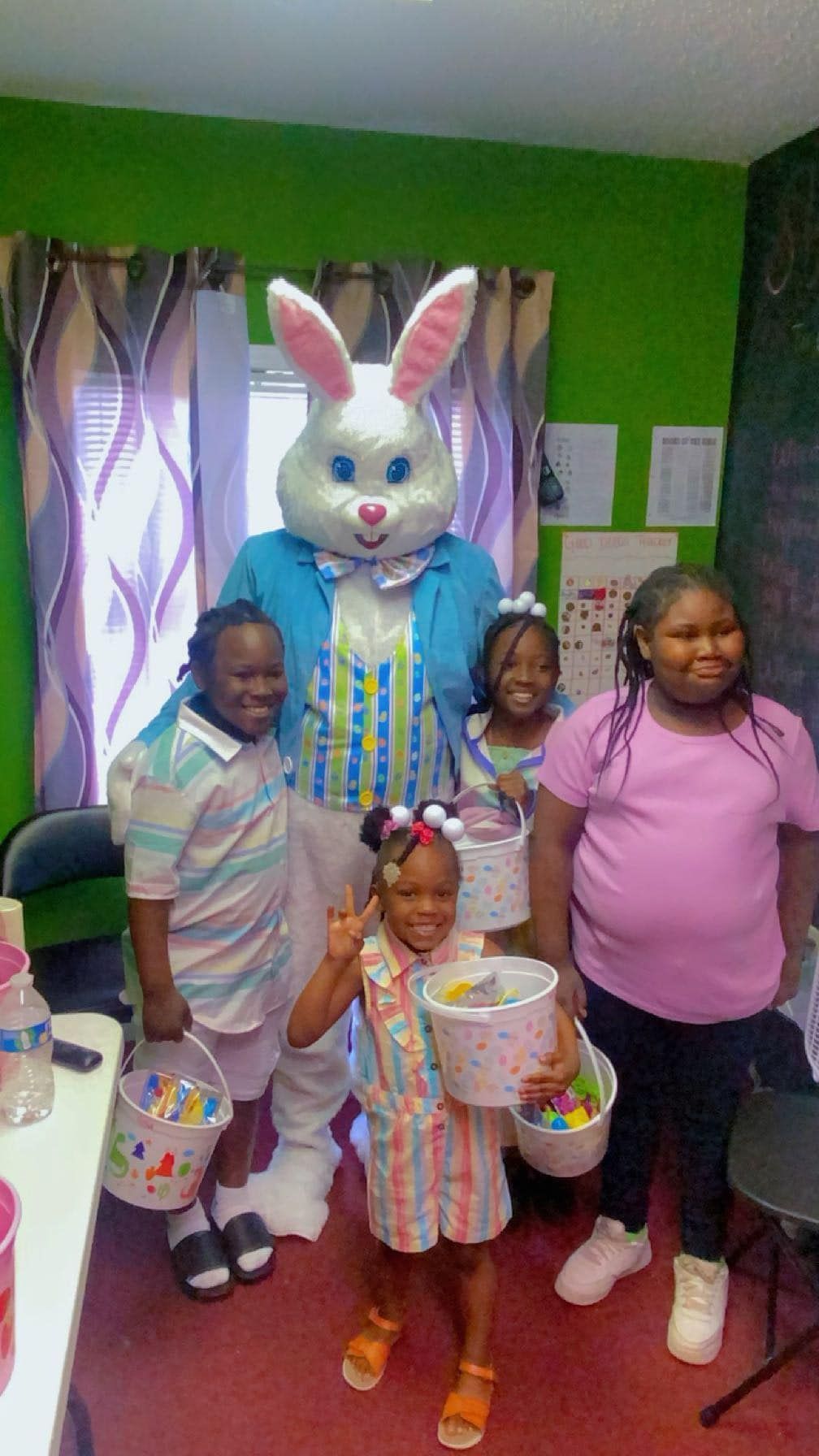 A group of four children stands in a brightly colored room with a person in an Easter bunny costume holding baskets.